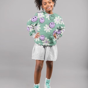 Può includere: Una persona giovane indossa una felpa verde con un motivo a margherite e faccine sorridenti, una gonna grigia e pattini a rotelle con ruote rosa. La felpa ha maniche grigie e i pattini hanno un design floreale.