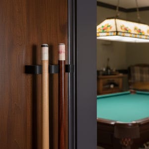 May include: A dark wood door with a black cue rack holding three pool cues. The cues are light brown and dark brown. In the background, a pool table with a green felt top is visible.