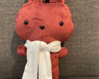 May include: A plush burgundy bunny toy wearing a white scarf. The bunny has black eyes and a simple, rounded design.