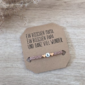 Könnte beinhalten: Ein pinkfarbenes Freundschaftsarmband mit goldenen Herzperlen und einem goldenen Buchstaben L-Anhänger. Das Armband liegt auf einer braunen Karte mit dem Text "Ein bisschen Mama, Ein bisschen Papa und ganz viel Wunder".