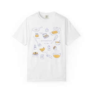 K&ouml;nnte beinhalten: Wei&szlig;es T-Shirt mit einer blauen Strichzeichnung von Fr&uuml;hst&uuml;ckszutaten, darunter eine Bratpfanne, Eier, Butter, Pfannkuchen und die Worte "breakfast club". Das Shirt hat einen Rundhalsausschnitt und kurze &Auml;rmel.