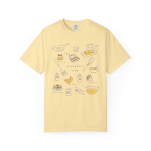 K&ouml;nnte beinhalten: Hellgelbes T-Shirt mit einem Fr&uuml;hst&uuml;cksmotiv. Das Design umfasst Illustrationen von Pfannkuchen, Eiern, Butter, Bananen und den Worten "Breakfast Club". Das Shirt ist aus einem weichen Material.