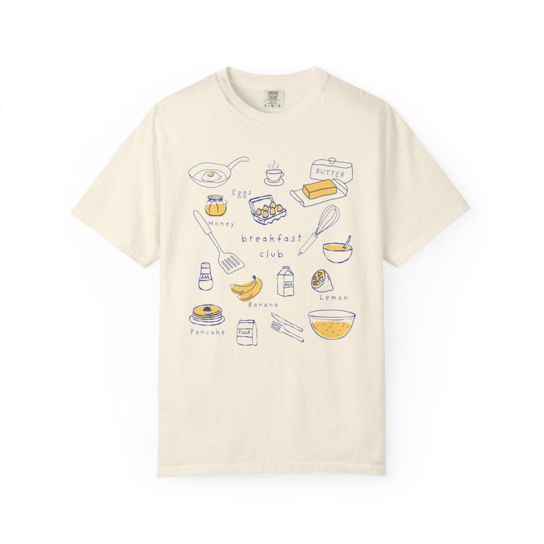 K&ouml;nnte beinhalten: Cremefarbenes T-Shirt mit einer blau-gelben Illustration von Fr&uuml;hst&uuml;cksartikeln. Das Design umfasst eine Bratpfanne, Eier, Butter, Honig, Pfannkuchen, Bananen und die Worte "breakfast club".