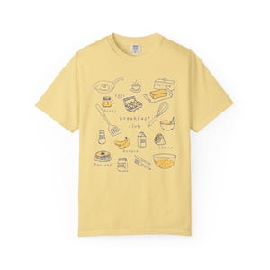 K&ouml;nnte beinhalten: Hellgelbes T-Shirt mit einer handgezeichneten Illustration zum Thema Fr&uuml;hst&uuml;ck. Das Design zeigt eine Bratpfanne, Eier, Butter, Honig, Pfannkuchen und den Schriftzug "breakfast club".