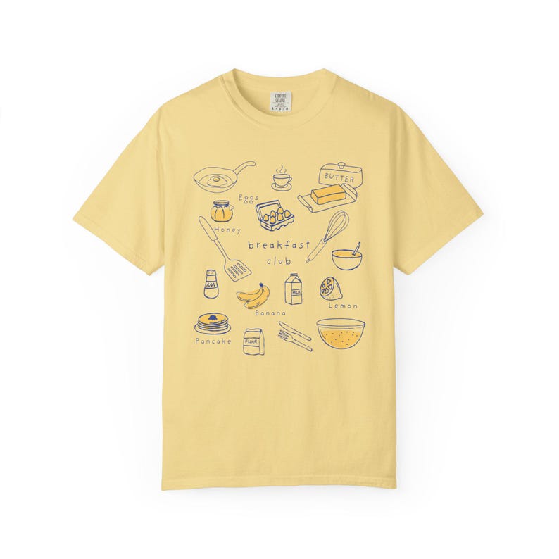 K&ouml;nnte beinhalten: Hellgelbes T-Shirt mit einer handgezeichneten Illustration zum Thema Fr&uuml;hst&uuml;ck. Das Design zeigt eine Bratpfanne, Eier, Butter, Honig, Pfannkuchen und den Schriftzug "breakfast club".