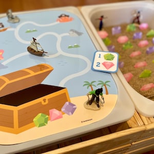 May include: A pirate-themed sensory play set with a treasure chest, map, toy ship, and gems. The set includes a sand-filled bin with colorful gems and small pirate figures. The map features a mermaid and a pirate island.