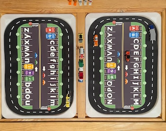 May include: Two black and white alphabet race tracks with toy cars and trucks on them. The tracks are on a wooden surface. The tracks have a stop sign at each end and arrows in the middle. The letters on the tracks are in alphabetical order.