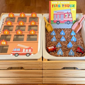 May include: A sensory bin set featuring a fire station board with numbered flame cutouts, a bin filled with brown beans, blue water drop cutouts numbered one through ten, two plastic scoops, two toy fire trucks, and a How It Works Fire Truck book.