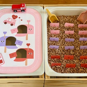 May include: A sensory play set with a Valentine's Day theme. The set includes a tray with a road design and mailboxes, a red toy truck, and a tray filled with beans and paper envelopes. Two scoops are included.