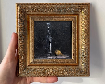 May include: A small square oil painting in an ornate gold frame. The artwork features a dark, moody background with a vertical white shape resembling a bottle and a small, textured yellow form resting at the base, rendered in thick paint.