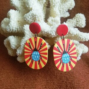May include: A pair of red and yellow earrings with a blue beetle design. The earrings are oval shaped and have a red and yellow striped pattern with a blue beetle in the center. The beetle has black legs and a blue body with white spots.