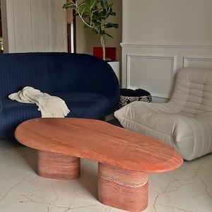 May include: A low-profile coffee table crafted from reddish-orange travertine stone, featuring an irregular oval top supported by two thick, cylindrical pedestal bases with a visible natural stone grain and a smooth, polished surface finish.