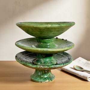 Op de afbeelding: Een groen keramisch serveerschaal met drie niveaus. De schaal heeft een voetstuk en drie cirkelvormige niveaus, elk met een iets andere diameter. Het groene glazuur heeft kleurvariaties, waardoor het een rustieke uitstraling heeft. Een lepel en een servet staan op de achtergrond.