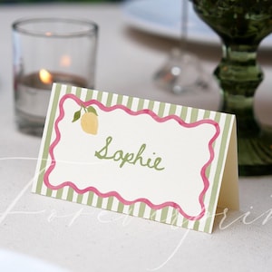 May include: A table place card featuring a lemon illustration and the name "Sophie" in green script. The card has a pink scalloped border and green and white striped background. A lit candle and green glass are in the background.