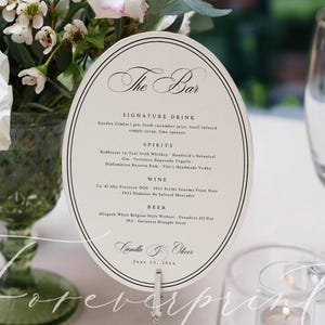 Puede incluir: Una tarjeta de menú ovalada con el texto "The Bar" y una lista de bebidas, colocada sobre una mesa con una copa de champán, un plato y una vela. Flores blancas en un jarrón verde en el fondo.