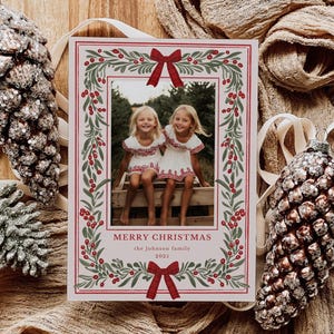 May include: Christmas card with a photograph of two smiling children, framed by a wreath of green leaves and red berries. The card has red bows and the text "MERRY CHRISTMAS" and "2025". Decorative pinecone ornaments are on either side.