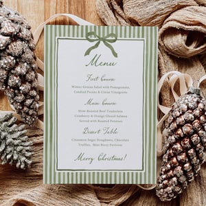 以下が含まれることがあります： 緑のストライプのボーダーとリボンのデザインが施された、お祝いのクリスマス メニュー カード。メニューには、前菜、メインコース、デザートテーブルが記載されています。装飾的な松ぼっくりのオーナメントとファブリックのアクセントが、お祝いのテーマを完成させています。