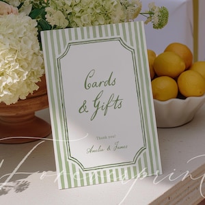 May include: A sign with green and white stripes that reads "Cards & Gifts" with the names Amelia & James. The sign is on a table with white wrapped gifts, lemons, and white flowers.