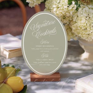 May include: An oval sign on a wooden base displays "Signature Cocktails" in elegant script. The sign lists two cocktail recipes: "Hers" and "His." A plate of lemons and a bouquet of white flowers are in the background.