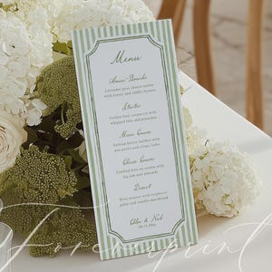 Op de afbeelding: Een menukaart met een groen-witte gestreepte rand, met een lijst van gerechten. Het menu staat op een witte tafel, omringd door witte bloemen en groen. Het menu bevat de namen van de gerechten en het bruidspaar.
