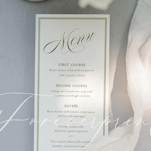 May include: A rectangular menu card with a green border and elegant script font. The menu lists courses like salad, soup, and entrees. A white fabric drapes across the image, adding a soft texture. The word "Menu" is at the top.