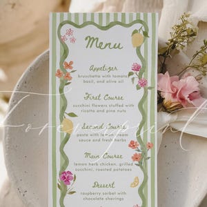 Può includere: Un biglietto del menu con un bordo a righe verdi e bianche, decorato con illustrazioni floreali e di limoni. Il menu elenca le opzioni di antipasto, primo piatto, secondo piatto, piatto principale e dessert. Il menu è su un piatto bianco con una composizione floreale.