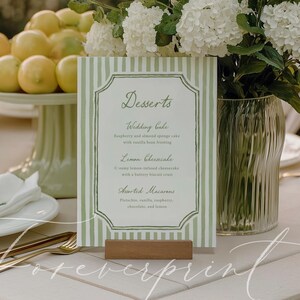 May include: A table setting with a menu card that reads "Desserts". The card is green and white striped with a wooden base. The table also has a cake stand with lemons, cupcakes, and a vase of white flowers.