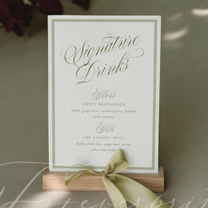 Puede incluir: Una tarjeta rectangular blanca con borde verde salvia que presenta el texto Signature Drinks, Hers Sweet Beginnings y His The Classic Mule, sostenida en un soporte de bloque de madera clara decorado con un lazo verde salvia.