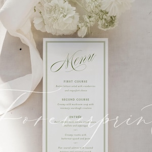 May include: A rectangular menu card with a green border and elegant script. The menu lists courses like butter lettuce salad, wild mushroom soup, basil chicken, and chocolate praline crunch cake. White flowers and a ribbon are in the background.