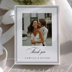 May include: A white wedding thank you card with a black border and the words "Thank you" in elegant script. The card features a photo of a smiling couple. A bouquet of white flowers and a white ribbon are in the background.