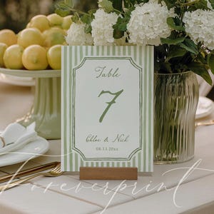 Puede incluir: Decoración de mesa de boda con un tarjetón con el número de mesa a rayas verdes y blancas. La tarjeta muestra "Mesa 7", los nombres "Chloe & Nick" y la fecha "08.13.20xx". Cubiertos dorados, platos blancos y un jarrón con flores blancas.