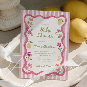May include: A baby shower invitation with a pink and white striped background. The invitation features floral and lemon illustrations, with text that reads "Baby Shower" and details for the event. Lemons and decorative elements surround the invitation.