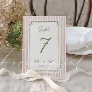 May include: A table number card with a pink and white striped background and a green border. The card displays the word "Table" and the number "7" in green script. The names "Chloe & Nich" and the date "08.13.20xx" are also printed on the card.