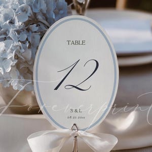 May include: An oval table number card featuring the text TABLE 12, S & L, and 08.21.20xx. The card has a light blue border and is held by a silver metal stand adorned with a white ribbon tied in a bow at the base of the card holder.