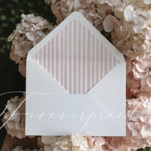 Könnte beinhalten: Ein weißer Umschlag mit rosa und weiß gestreiftem Innenfutter, geöffnet, um das Muster zu zeigen. Der Umschlag ist von hellrosa Blüten umgeben. Das Wort "foreverprint" ist kursiv geschrieben.