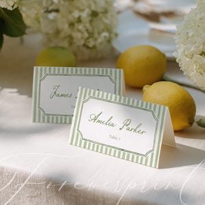 May include: Two place cards with green striped borders and elegant script names, alongside lemons and white flowers. The cards read "James" and "Amelia Parker, Table 1". The scene is set on a white tablecloth.