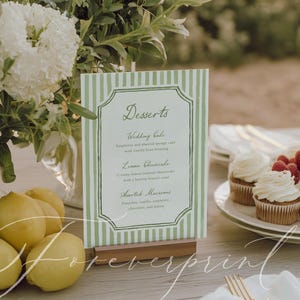 May include: A table setting featuring a green and white striped menu card with the word "Desserts". The menu is in front of a bouquet of white flowers, lemons, and cupcakes with raspberries. The menu is held in a wooden stand.