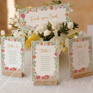 May include: A set of rectangular paper seating chart cards with floral borders in shades of pink, orange, and green. The cards feature gold text reading Find your Seat, Table 1, Table 2, and Table 3, each held upright by small wooden blocks.