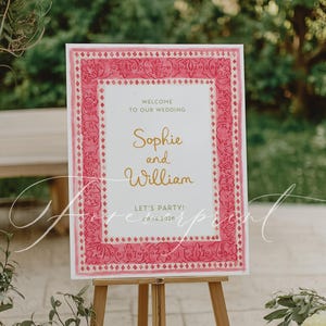 May include: A white sign on a wooden easel with a pink floral border. The sign reads "Welcome to our wedding" and features the names "Sophie and William" in gold script, with the date "09.14.2026" below.
