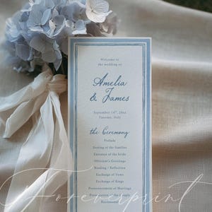 Op de afbeelding: Een trouwprogramma met een lichtblauwe rand. Het programma bevat de namen "Amelia & James" en de datum en locatie van de bruiloft. Een boeket blauwe hortensia's en wit lint staan op de achtergrond.