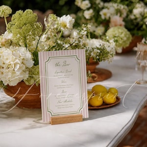 May include: A bar menu card with pink and white stripes, displayed on a wooden stand. The card features the text "The Bar" and a list of drinks. The scene includes floral arrangements and lemons.