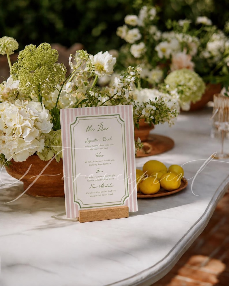 May include: A bar menu card with pink and white stripes, displayed on a wooden stand. The card features the text "The Bar" and a list of drinks. The scene includes floral arrangements and lemons.