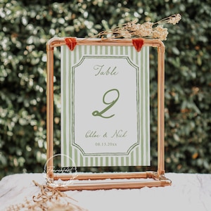 May include: A copper-colored metal table number holder with a green and white striped card. The card displays the word "Table", the number "2", and the names "Chloe & Nick" with a date. Dried flowers and red velvet accents adorn the top.