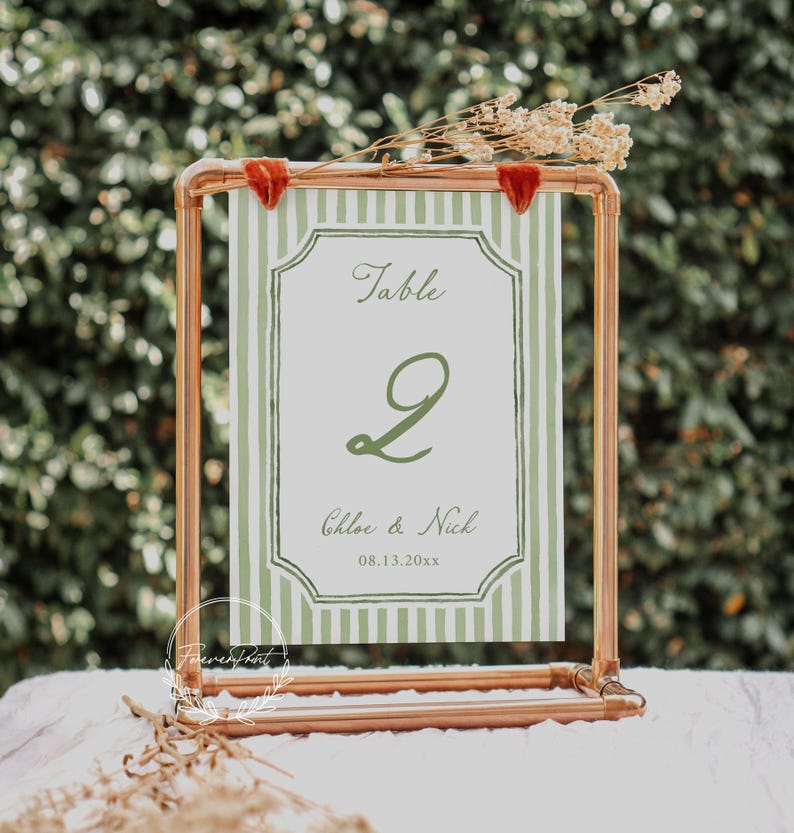 May include: A copper-colored metal table number holder with a green and white striped card. The card displays the word "Table", the number "2", and the names "Chloe & Nick" with a date. Dried flowers and red velvet accents adorn the top.