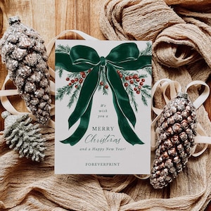 Può includere: Un biglietto di auguri natalizio con un fiocco verde e la scritta "Buon Natale e Felice Anno Nuovo!" è circondato da pigne decorative e tessuto. Il biglietto presenta un design festivo con bacche rosse e rami di sempreverdi.