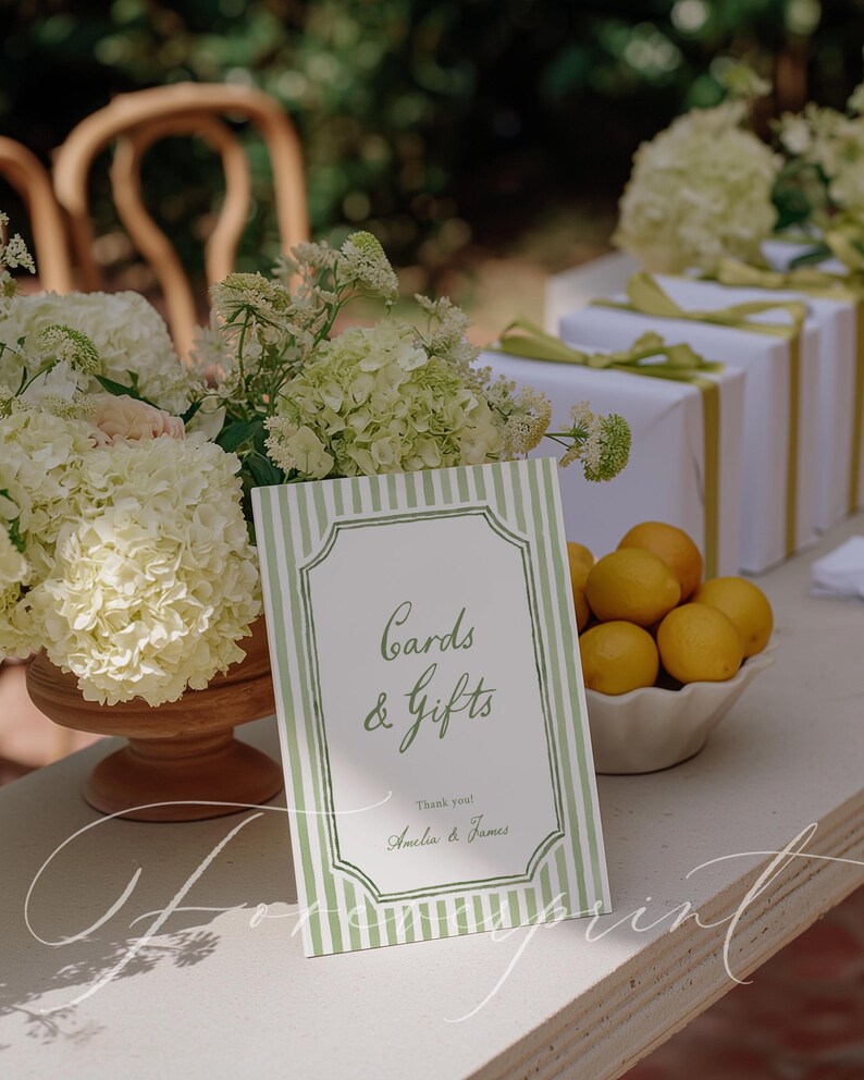 May include: A sign with green and white stripes that reads "Cards & Gifts" with the names Amelia & James. The sign is on a table with white wrapped gifts, lemons, and white flowers.