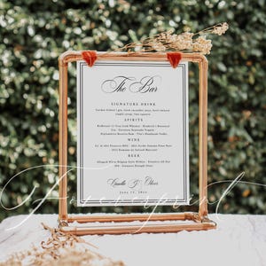 May include: A copper-coloured metal framed sign with a white insert, displaying a bar menu. The menu lists signature drinks, spirits, wine, and beer. The sign is decorated with dried flowers and red velvet accents.