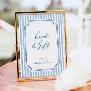Pu&ograve; includere: Un cartello con cornice dorata con sfondo a righe blu e bianche e le parole "Cards & Gifts" in elegante scrittura. Il cartello include anche il testo "Thank you! Amelia & James."