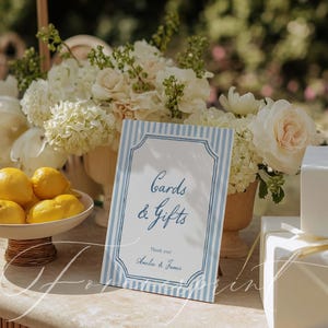 Pu&ograve; includere: Un cartello a righe blu e bianche con la scritta "Cards & Gifts" e i nomi "Amelia & James" in basso. Il cartello &egrave; su un tavolo con fiori bianchi, limoni e regali confezionati.