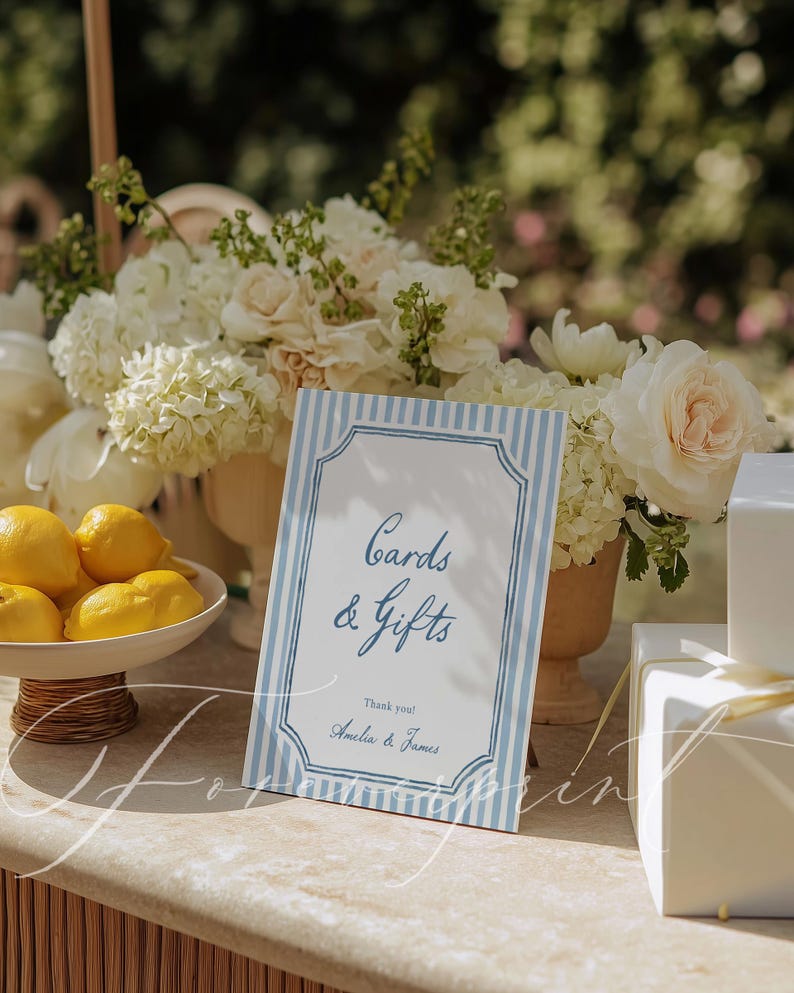 Pu&ograve; includere: Un cartello a righe blu e bianche con la scritta "Cards & Gifts" e i nomi "Amelia & James" in basso. Il cartello &egrave; su un tavolo con fiori bianchi, limoni e regali confezionati.